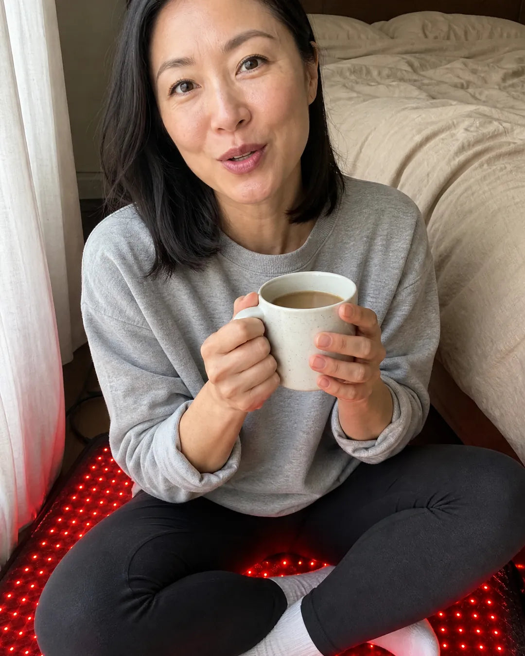 Woman sitting cross-legged on the EMBRA red light mat on her bedroom floor with a cup of coffee